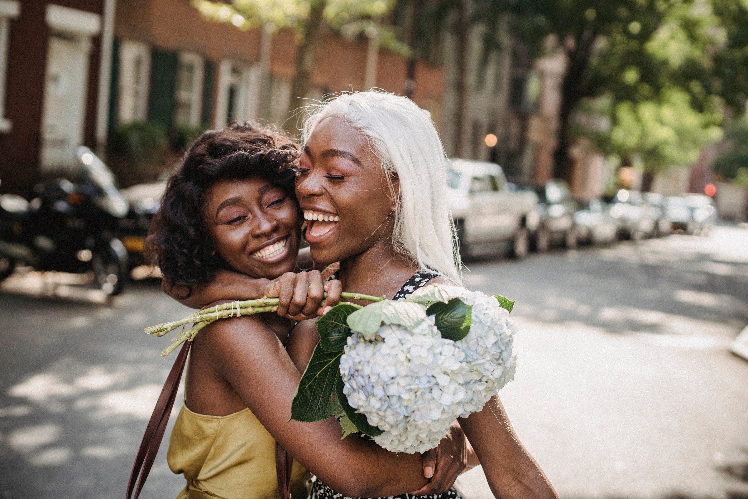 Two women laughing and embracing on a sunny city street, one holding a bouquet of white hydrangeas.